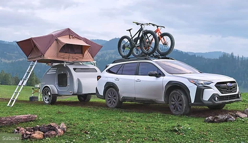 Subaru Outback Pulling a Small, Silver Teardrop Camper With a Roof-Mounted Foldable Tent, Parked on a Grassy Field With a Backdrop of a Forest and Hilly Landscape