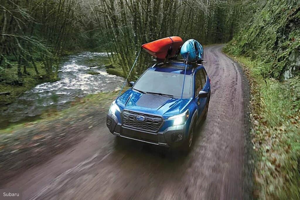A blue Subaru Forester with kayaks on the roof rack is driving along a curvy forest road next to a stream. The car is in motion, which is captured by the slight blur of the trees and the road. One kayak is red and the other is turquoise, adding a pop of color to the scene. The setting is lush and green, suggesting a natural, possibly mountainous location ideal for outdoor activities.