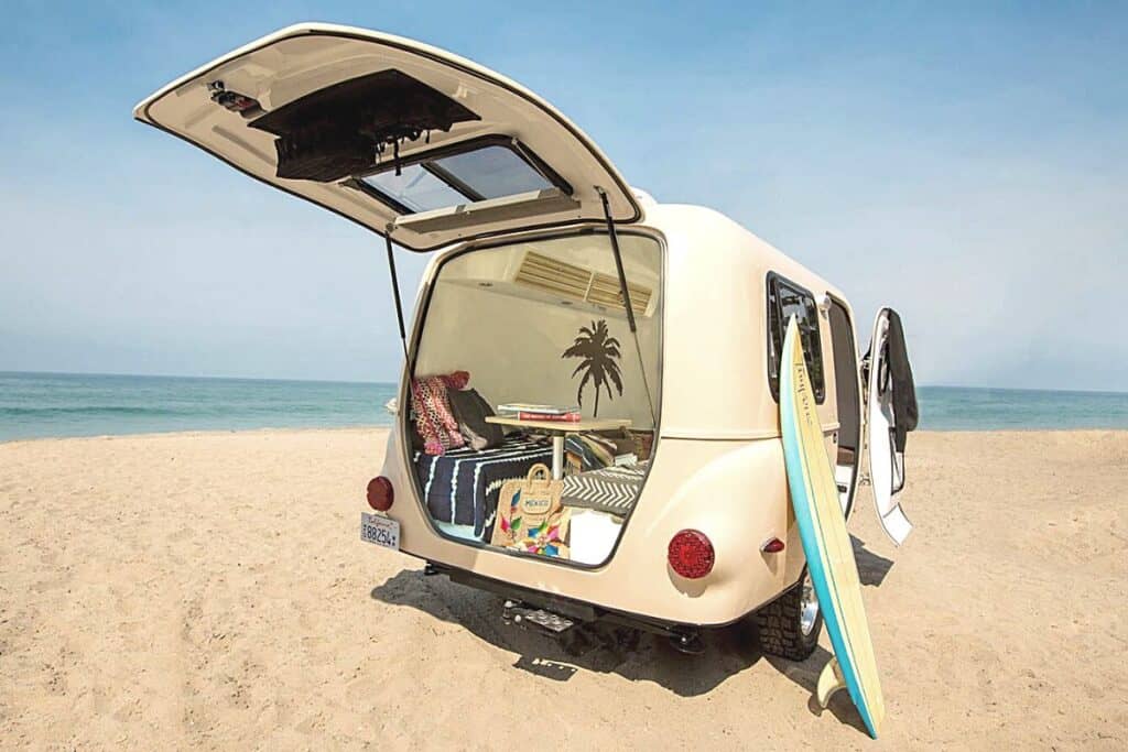 A Happier Camper HC1 is positioned on a sandy beach with the ocean in the background. The rear hatch is open, displaying a colorful and inviting interior packed with beach essentials, including towels, a tote bag, and a surfboard leaning against the camper. The interior features a cushioned seating area with a palm tree design on the window. The camper's cream-colored exterior is complemented by the clear blue sky, and both doors are wide open, inviting in the seaside air.
