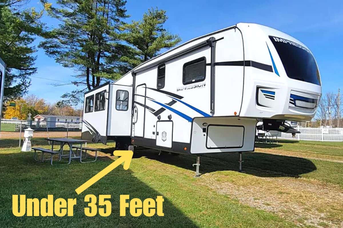 A 5th wheel parked outdoors with "Under 35 Feet" labeled, indicating its length. The RV features a modern design with a blue and black graphic on a white background, a prominent over-cab area, and multiple slide-outs. It's a sunny day, and the RV is situated on a grassy area with a picnic table to the side, trees in the background, and a clear blue sky above.