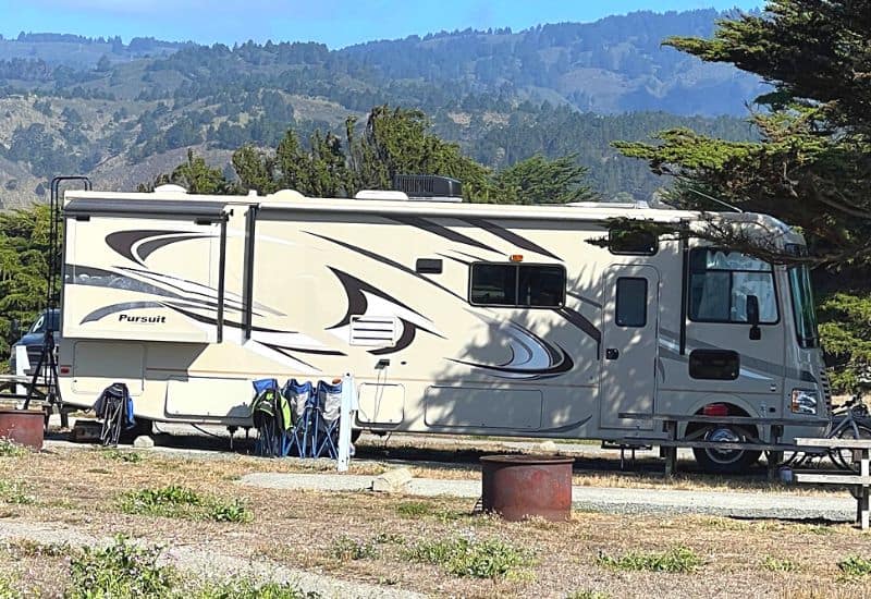 Beige Class A "Pursuit" Motorhome Parked at a Campsite With a Backdrop of Lush Green Hills and Trees Under a Clear Blue Sky