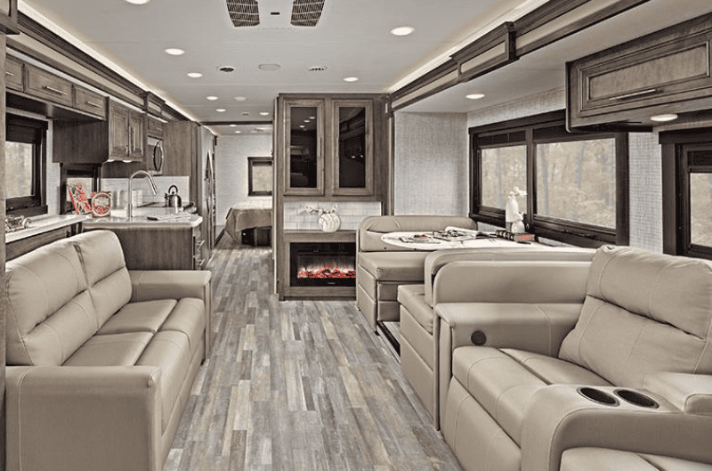 Well-lit Interior View of a Class A Motorhome Featuring a Cozy Living Space With Two Beige Couches, a Booth Dinette, a Fireplace, and a Kitchen in the Background
