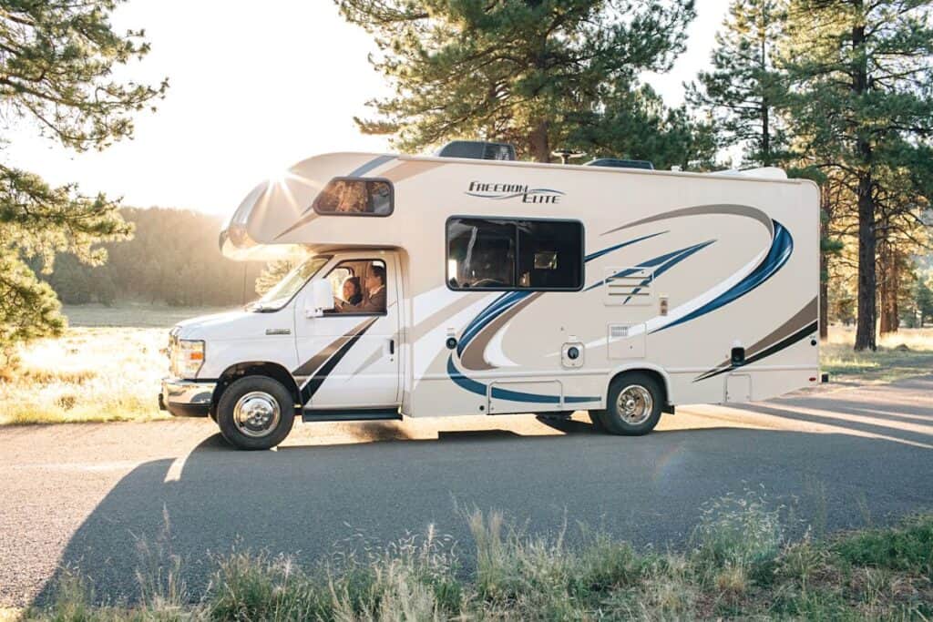 Class C Motorhome Driving on an Asphalt Road in a Pine Forest with the Sun Shining Brightly from Behind, Casting a Warm Glow and Creating Lens Flare