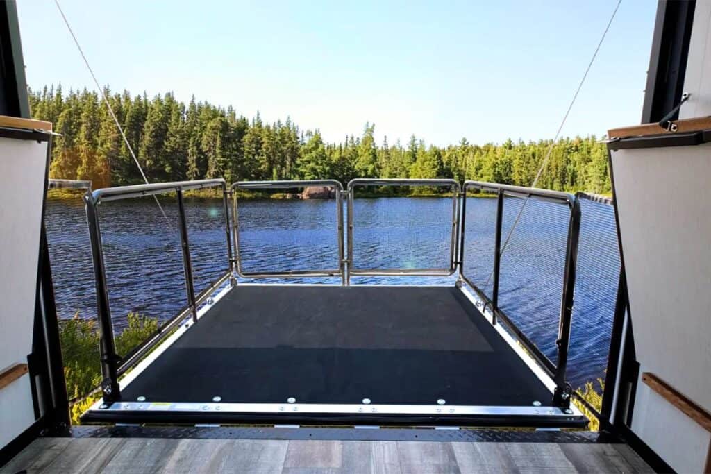 Open rear ramp of the Forest River Wolf Pup 18RJBW toy hauler, converted into a patio deck overlooking a serene lake surrounded by a forest.