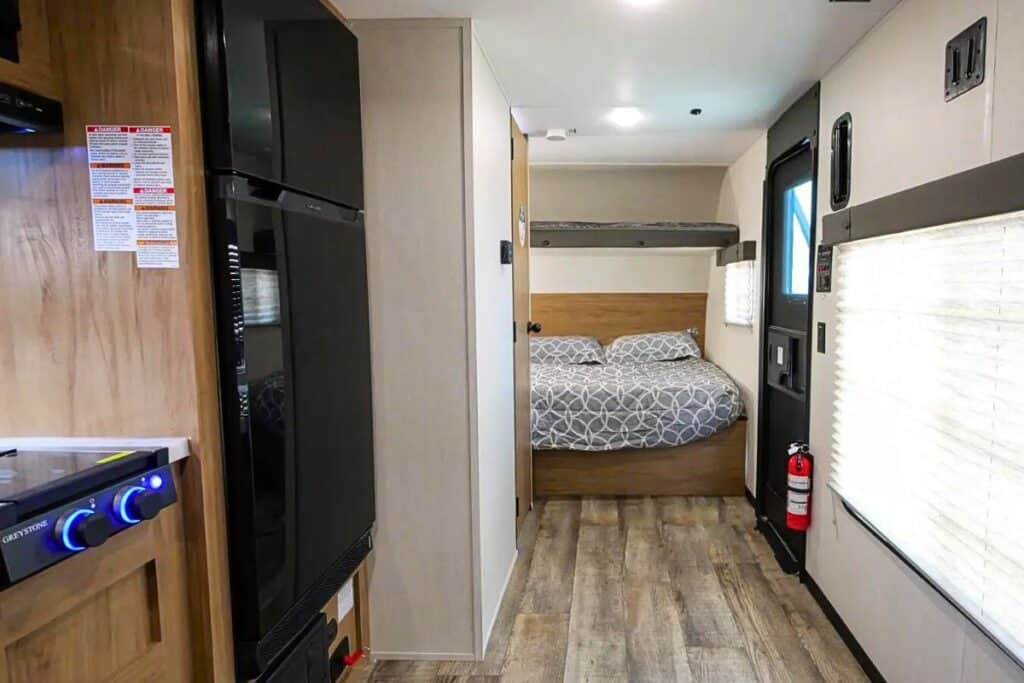 Inside the Forest River Wolf Pup 18RJBW toy hauler. On the left, there is a kitchen area featuring a tall, slim refrigerator in black, with wooden cabinet doors above and below it. In the foreground, a cozy sleeping area is equipped with a queen bed dressed in patterned bedding. Above the bed, a bunk provides additional sleeping space. Natural light streams in through the windows, which are covered with blinds.