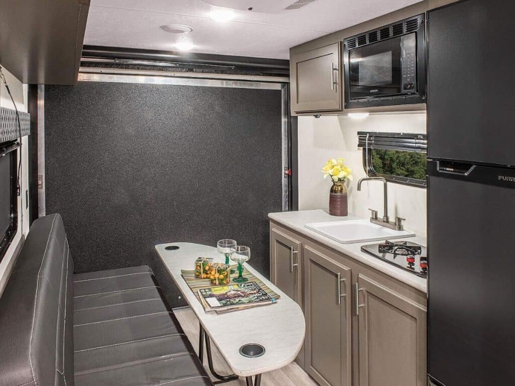 Inside the KZ RV Sportsmen Classic 180TH, there is a cozy dining area with booth-style seating and a table, next to a compact kitchen with a sink, stove, microwave, and refrigerator, all accented with wooden cabinetry.