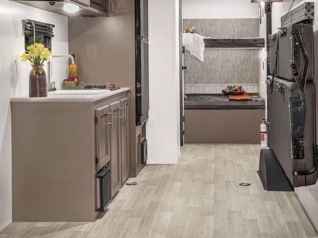 Interior view of the KZ RV Sportsmen Classic 180TH toy hauler showing a compact kitchenette with a sink and stove on the left, and an opposite wall featuring a leather-strapped fold-down sofa bed in an upright stored position.