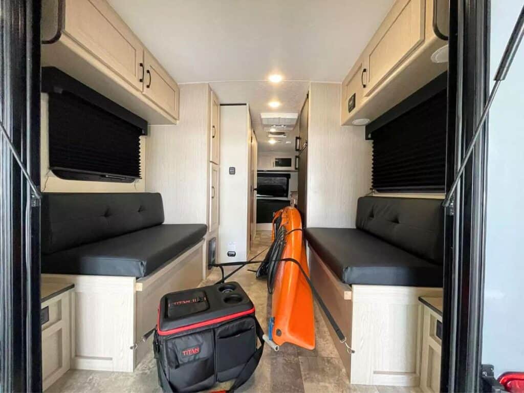 The interior of the Coachmen Apex Nano 17TH viewed from the rear hatch, which features side bench seating with storage underneath, overhead cabinets, closed blinds, and a central walkway leading to the front of the toy hauler, with an orange kayak and a black and red bag on the floor.