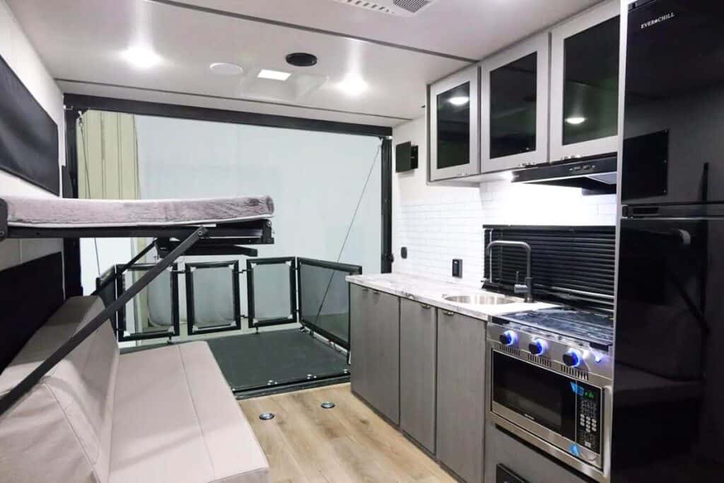 Inside the Forest River Ozark 1900THK toy hauler, looking towards the rear ramp door, adjacent to the kitchen featuring modern appliances, including a stove with blue-lit knobs, and overhead cabinets, with an elevated bunk bed to the left above a beige sofa.