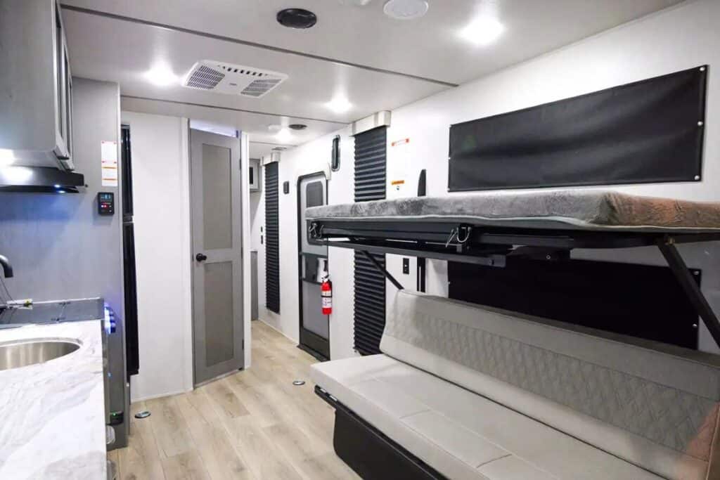 Inside the Forest River Ozark 1900THK, showcasing a sleek kitchen on the left, a corridor leading to the entry door, and on the right, a sofa with a foldable bunk bed above it, set against a backdrop of contemporary decor and wood-look flooring.
