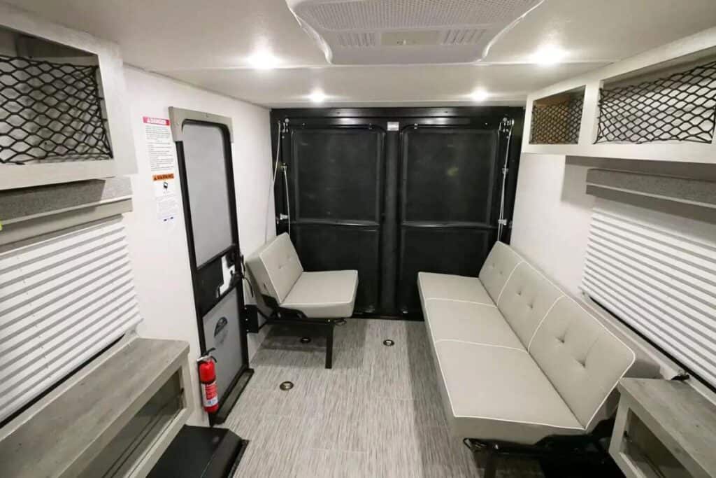 The Interior of the Forest River Ibex 19QTH toy hauler showing facing bench seats, overhead net storage, and the rear door closed, with a fire extinguisher visible to the left and wood-look flooring throughout.
