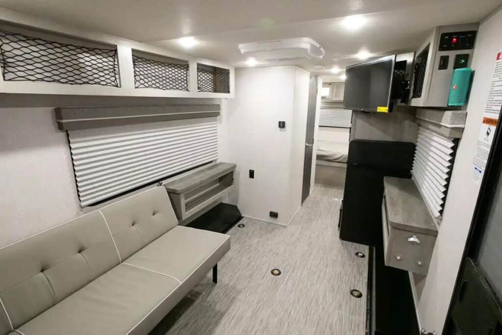 Interior of a Forest River Ibex 19QTH toy hauler showing a convertible sofa bed, overhead net storage, a central aisle leading to a bedroom, and a compact storage area with a TV on the right.