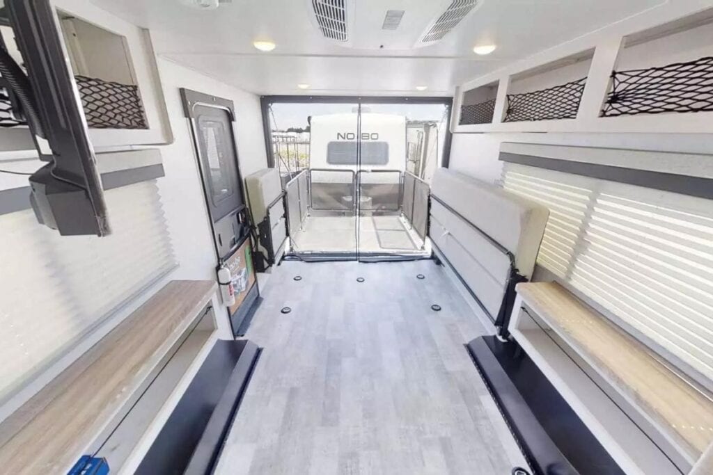 Interior of the Forest River No Boundaries NB19.1 toy hauler, featuring a bright and airy space with bench seating along the walls, overhead net storage, and an open rear door leading to an outdoor patio area, all under a well-lit ceiling with recessed lights.