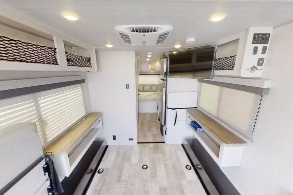 The interior of the Forest River No Boundaries NB19.1 toy hauler, featuring parallel cabinets with overhead net storage, a central walkway leading to a compact kitchen area on the right with a refrigerator, and a bedroom area visible in the back, all illuminated by ceiling lights.