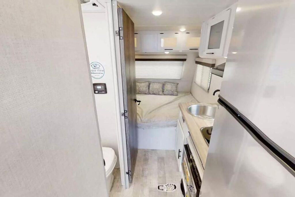 The interior view towards the front of the Forest River No Boundaries NB19.1 toy hauler, showing a bathroom on the left, a cozy bedroom area with a window and overhead cabinets straight ahead, and a kitchen countertop with a sink on the right.