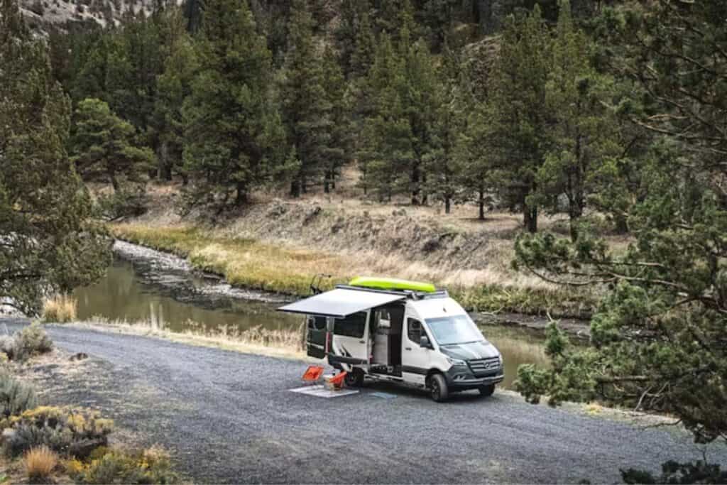 A Class B RV parked by a serene river surrounded by forested hills.